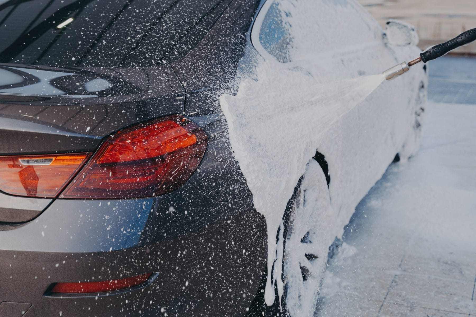Car exterior cleaning. Applying snow foam on dirty auto surface from high-pressure washer at carwash station outdoors, worker spraying suds on auto with special sprayer