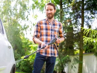 Car washing in the garden with a high pressure washer on a warm sunny day. A young bearded man using a compact home high pressure washer. Backyard, exterior, outdoor car cleaning.