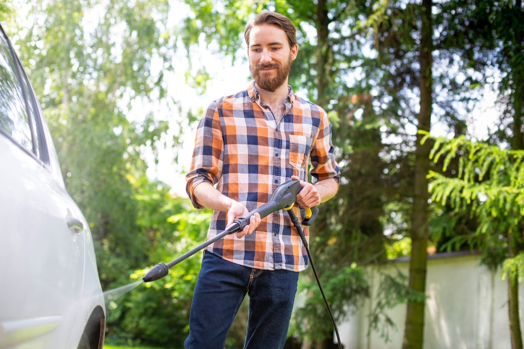 Car washing in the garden with a high pressure washer on a warm sunny day. A young bearded man using a compact home high pressure washer. Backyard, exterior, outdoor car cleaning.