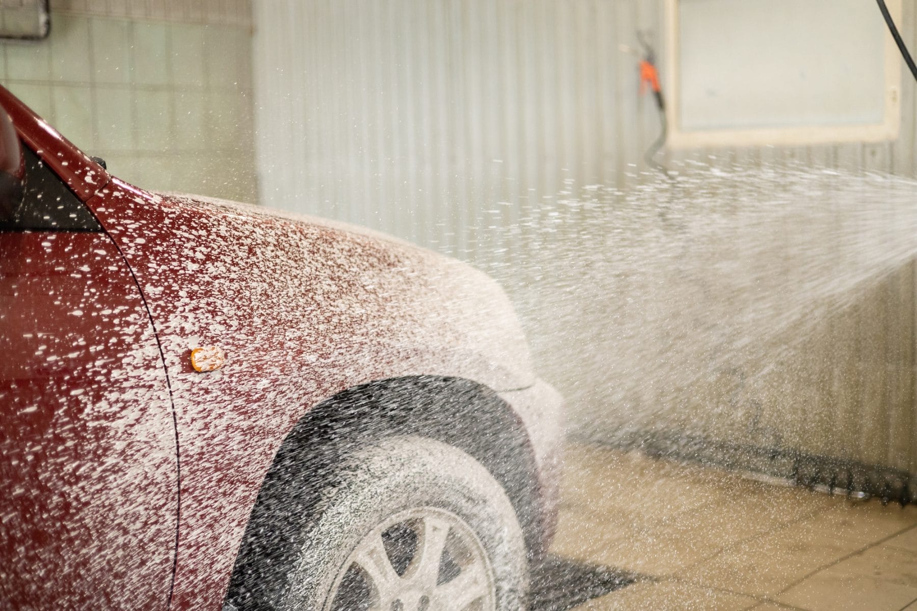 At a touchless car wash, the driver applies foam from a gun. The red car is covered with soap suds in the box of the atomic washer. The man drove his car to the car wash to wash off the dirt and dust.